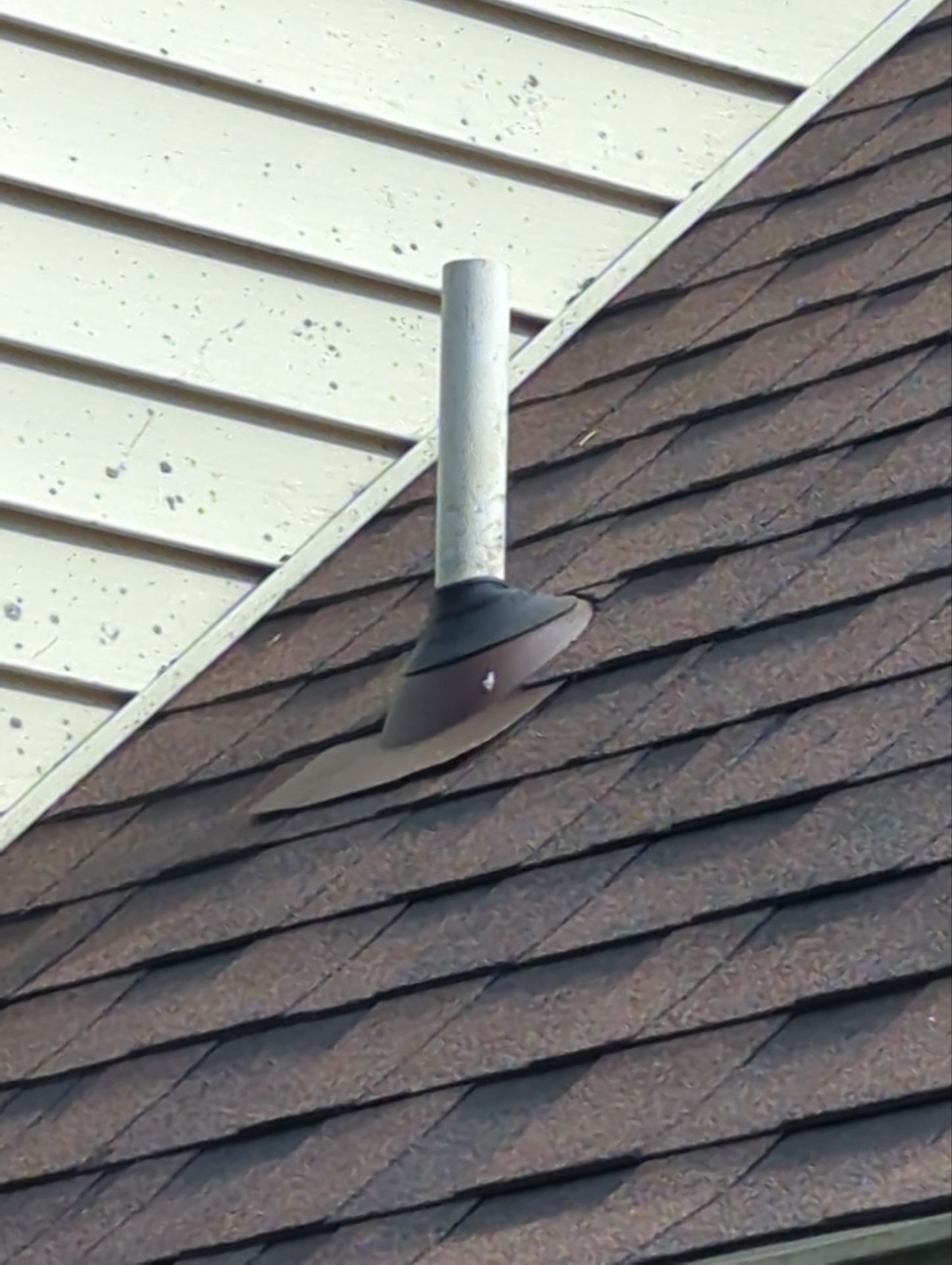 Metal vent pipe on brown shingled roof, next to cream-colored siding.