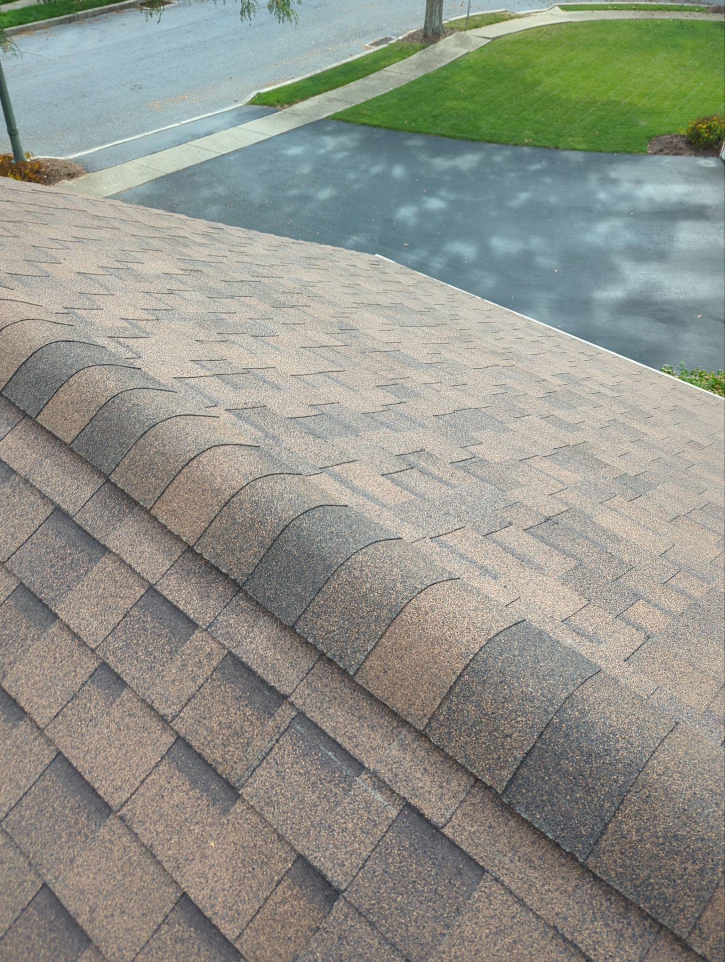 View of a brown and gray shingle roof from above with a paved driveway and lawn in the background.