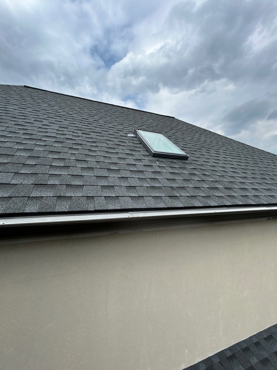 Gray shingle roof with a skylight, against a cloudy sky.
