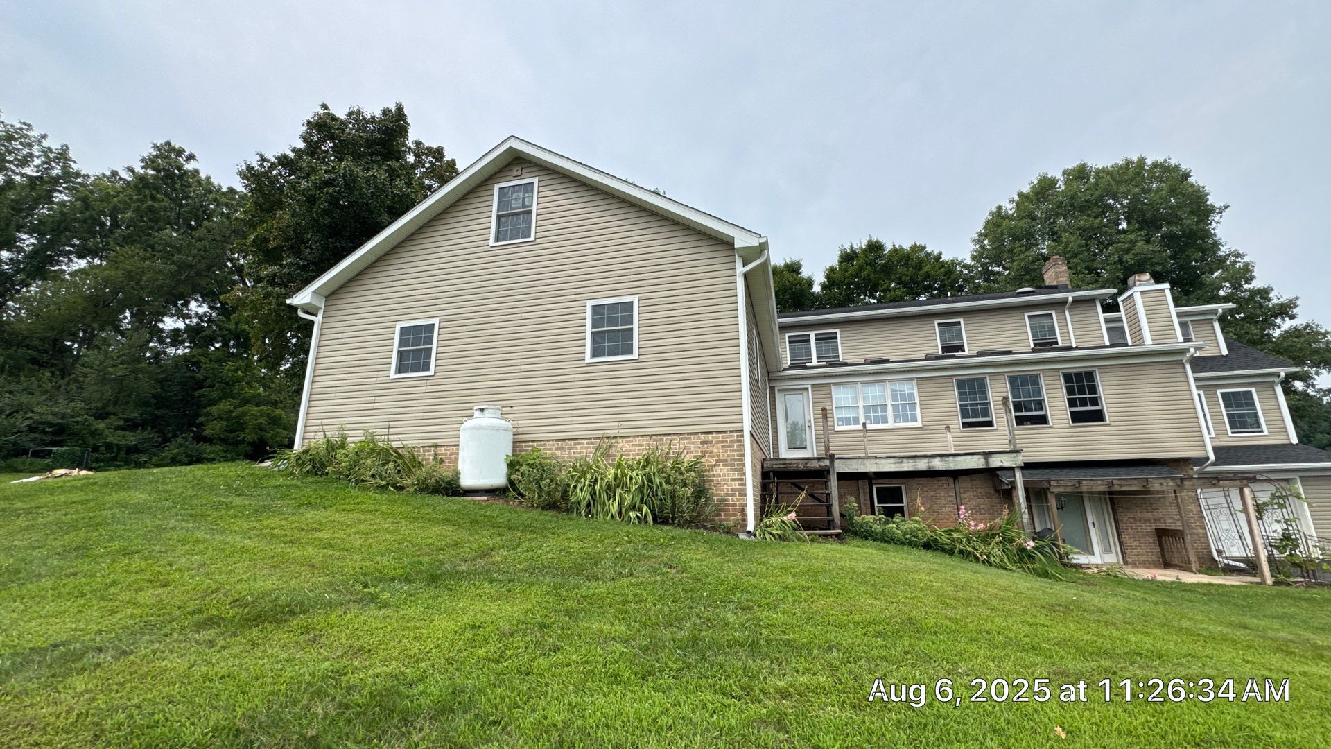 Tan house with green grass on a hillside. Trees in the background. White propane tank on the side of the house.