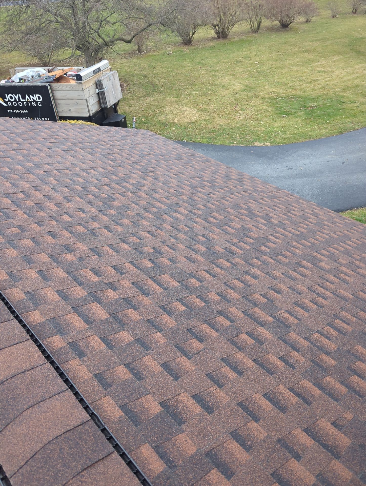 Brown asphalt shingle roof on a house, with a truck in the background. Green grass and driveway are also visible.