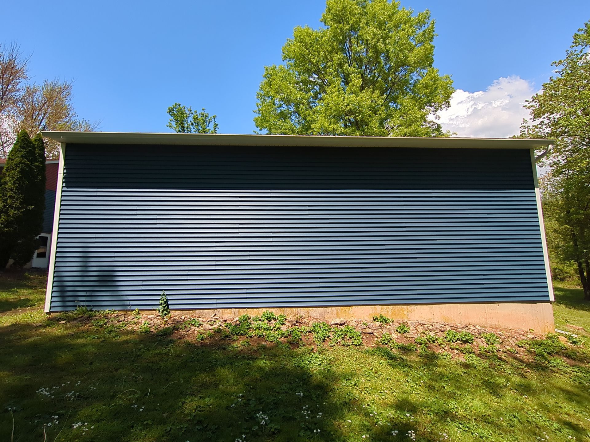 Blue corrugated metal shed with a flat roof, set on a grassy lawn with trees and a blue sky in the background.