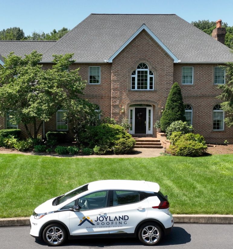 A white Joyland Roofing car parked in front of a brick house with a newly installed gray roof.