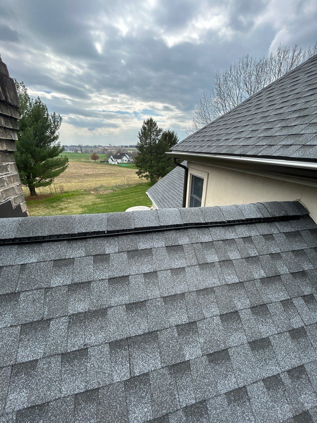 Gray asphalt shingle roof on a cloudy day with a view of a green field and distant buildings.