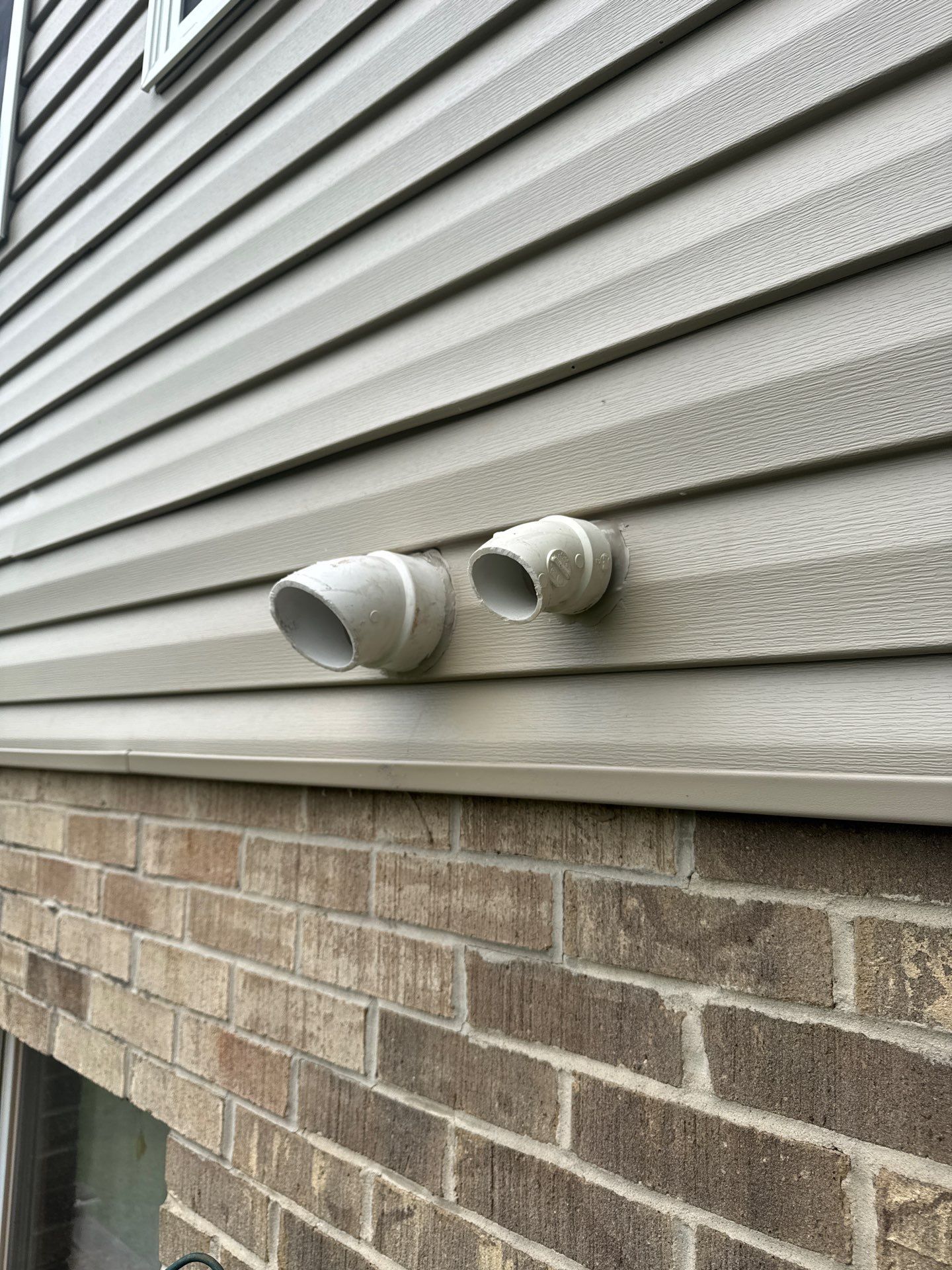 Two white vents on tan siding above a brick foundation.