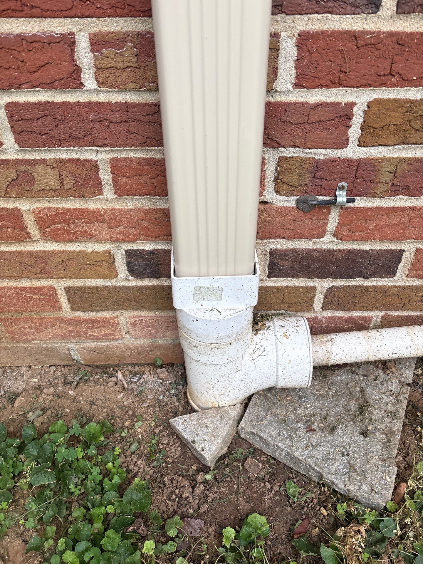 Beige gutter and downspout against a brick wall, resting on concrete blocks and directing water away.