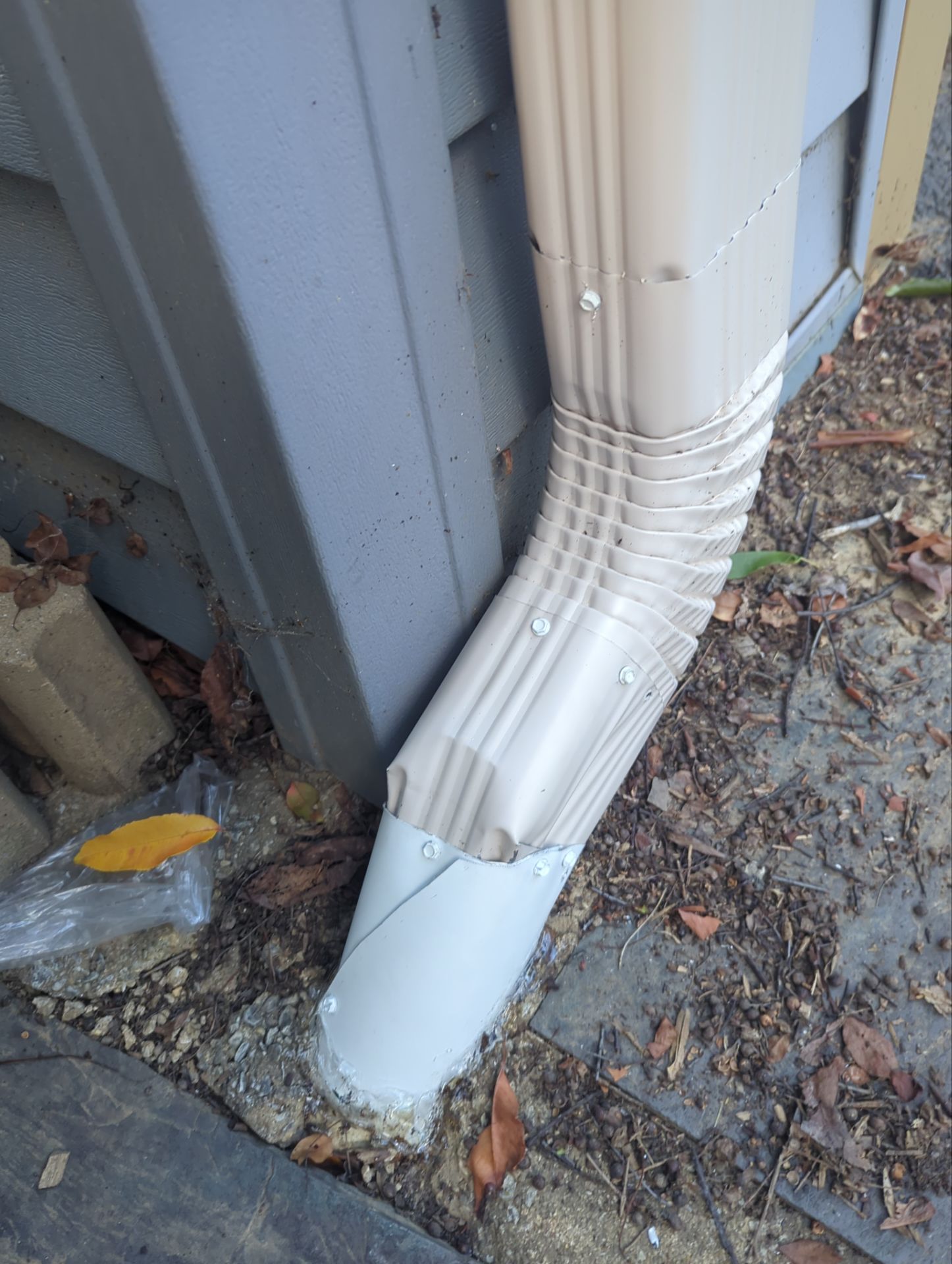 Beige gutter downspout attached to a building, angled toward the ground and debris.