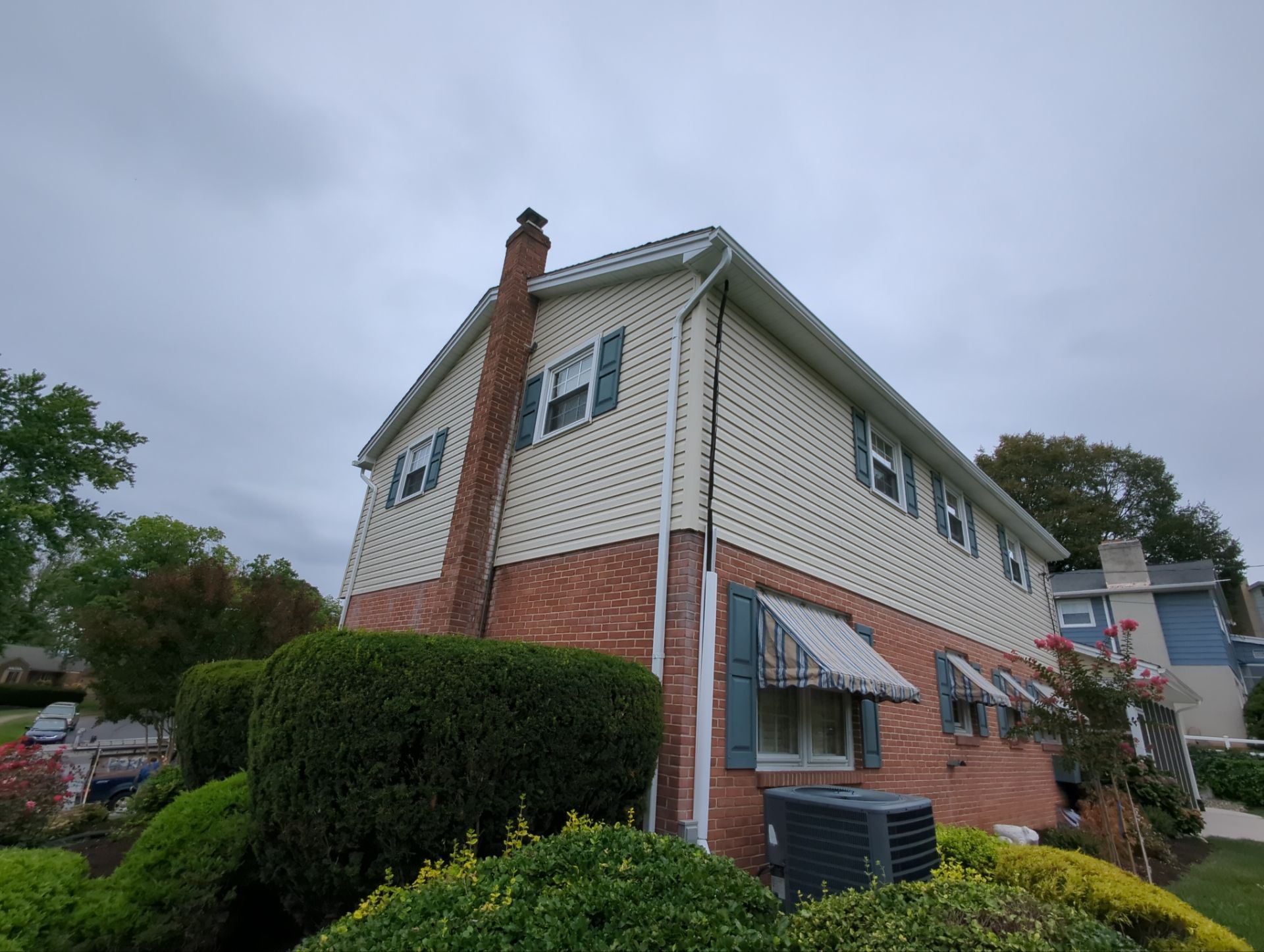 Two-story house with tan siding, black shutters, white porch with hanging plant, and a cloudy sky.