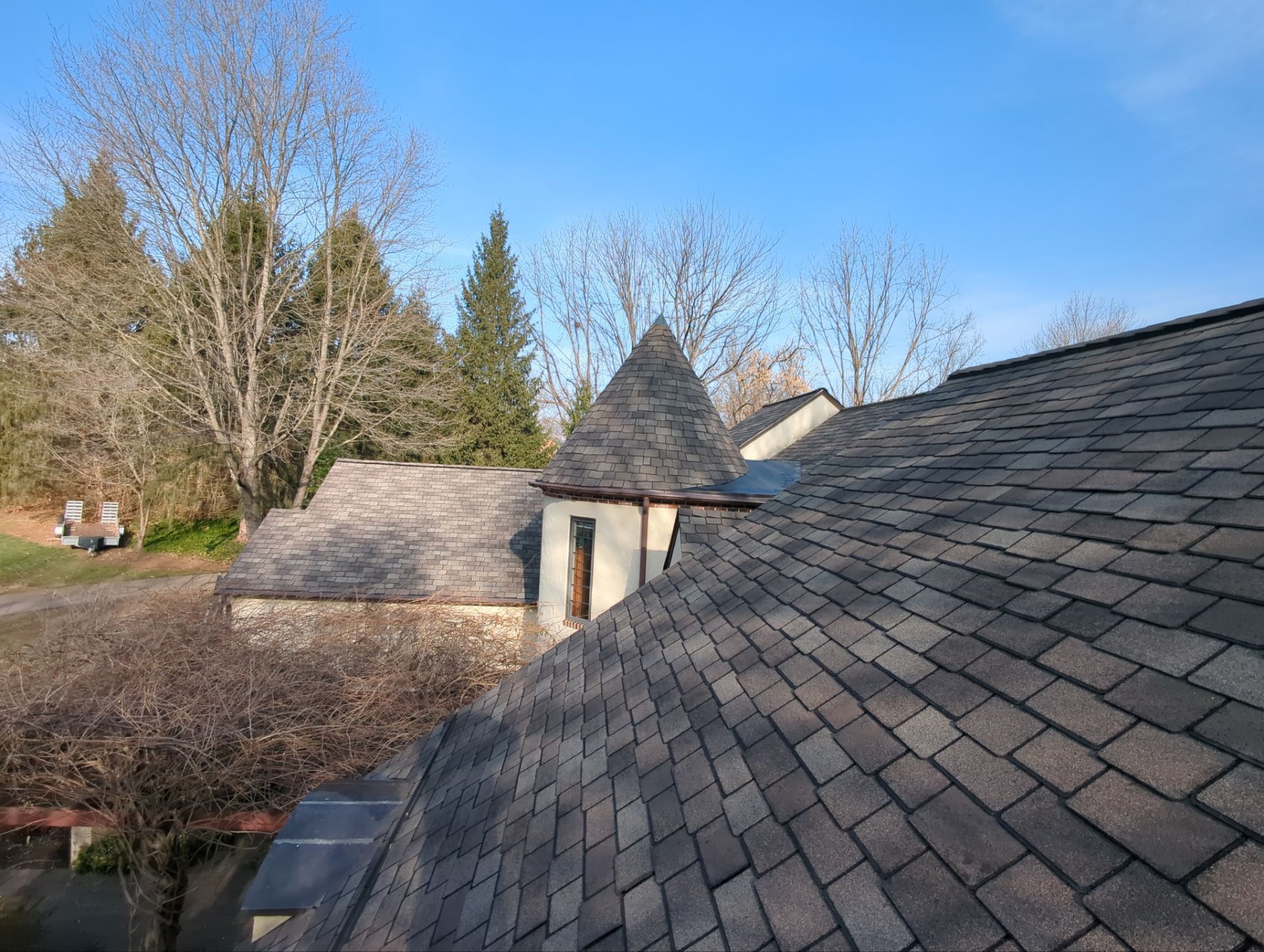 Asphalt shingle roof on a house with a small turret. Bare trees and blue sky in background.