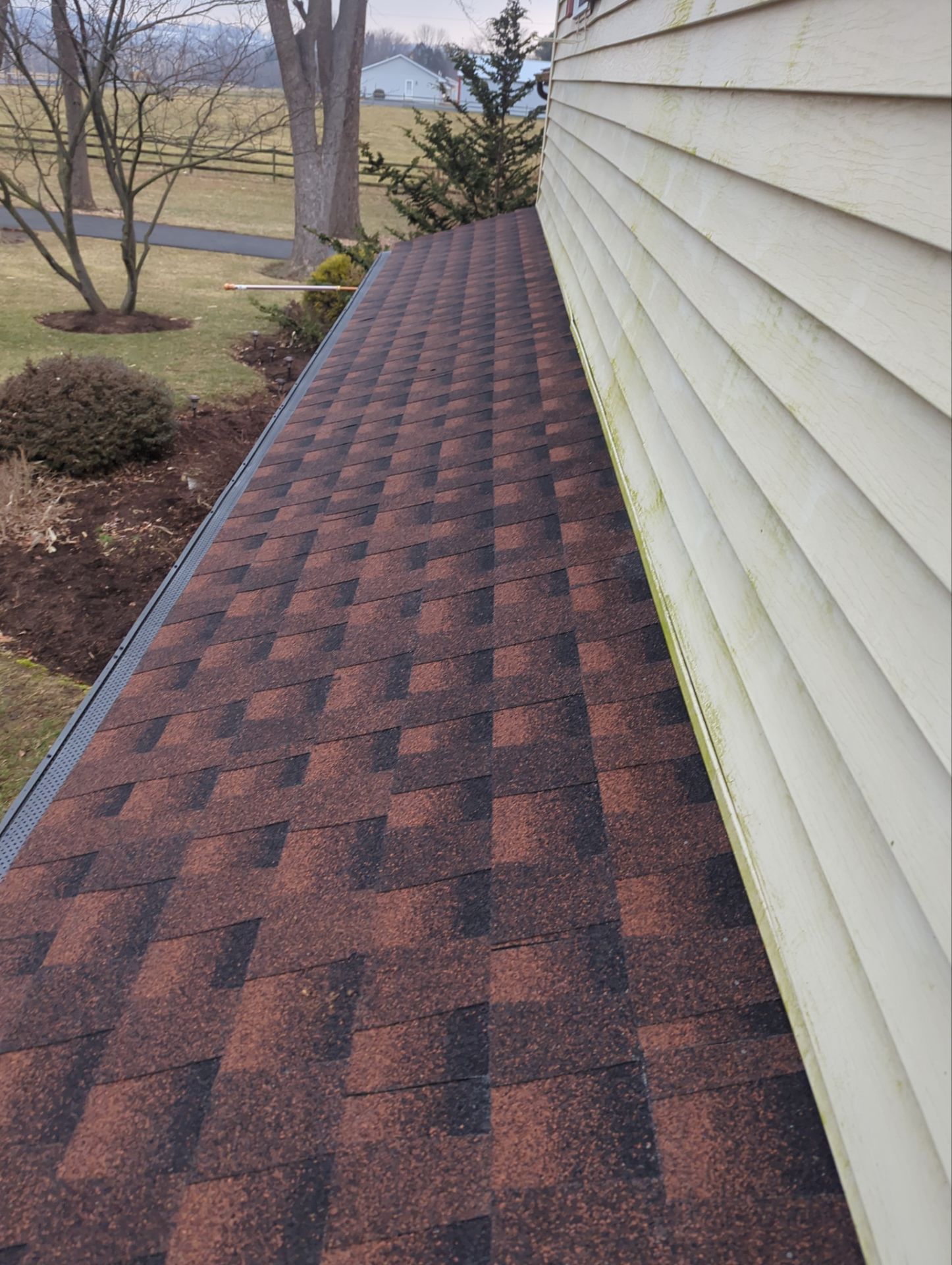Dark brown and black shingle roof adjacent to light yellow siding on a house. Green grass and trees visible.