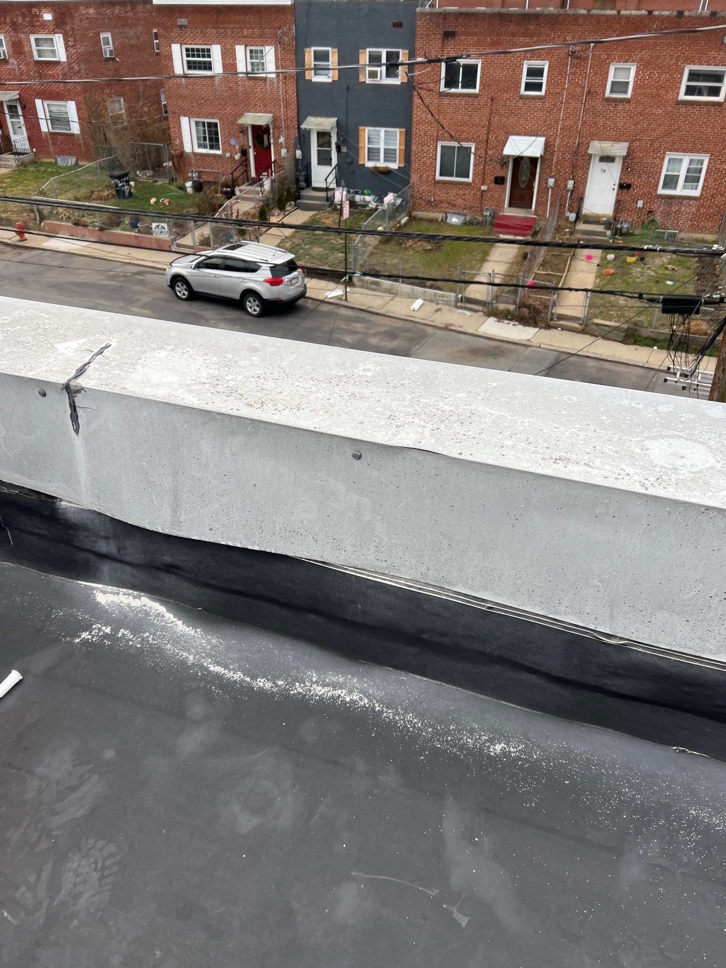 View from a rooftop: concrete barrier and asphalt roof, overlooking street and row houses; car parked on street.