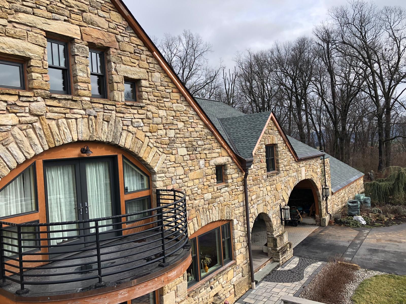 Stone house with arched entryway, balcony, and garage; trees in background.