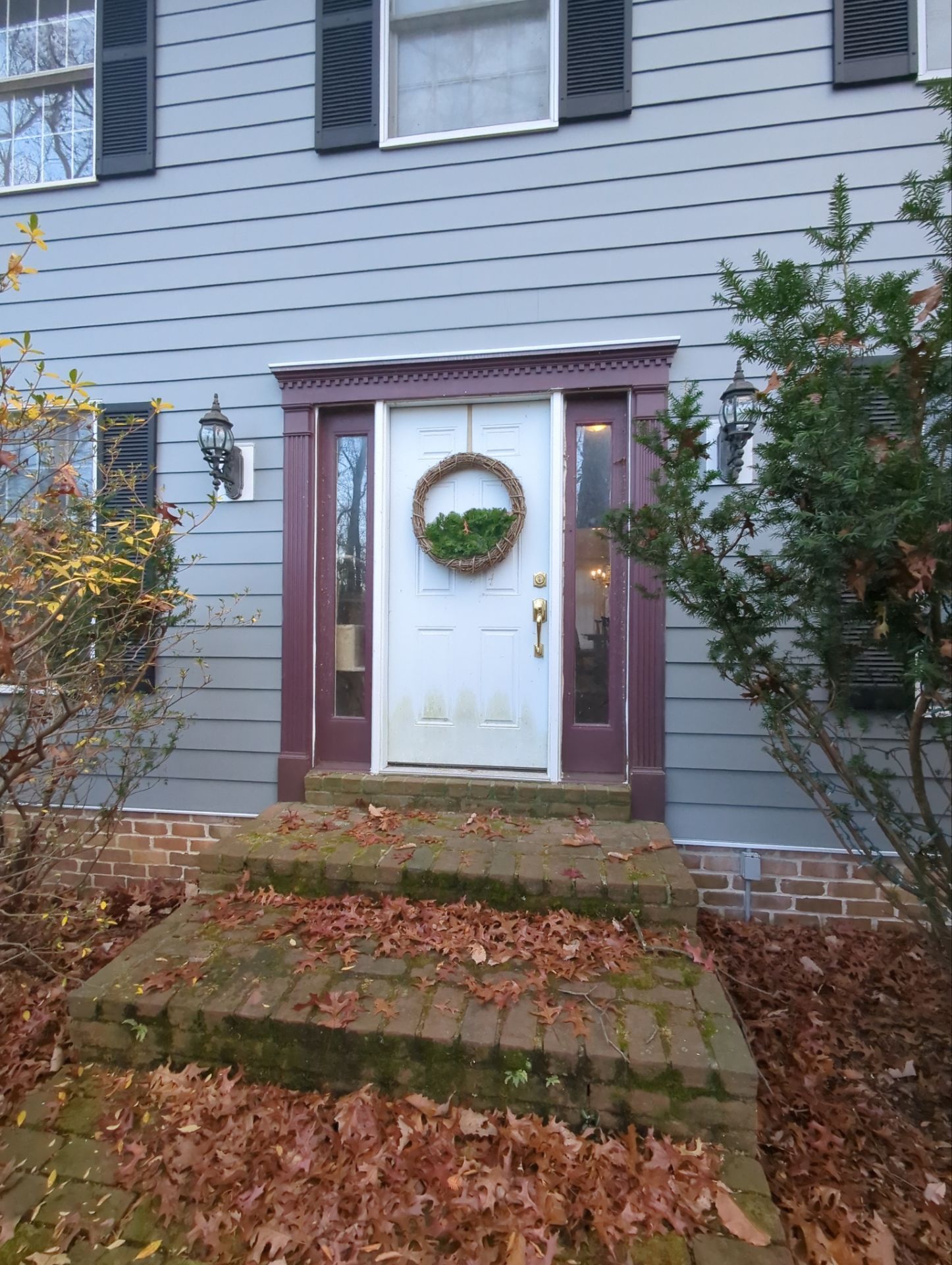 Grey house front with red-trimmed door, wreath, and brick steps covered in fallen leaves.