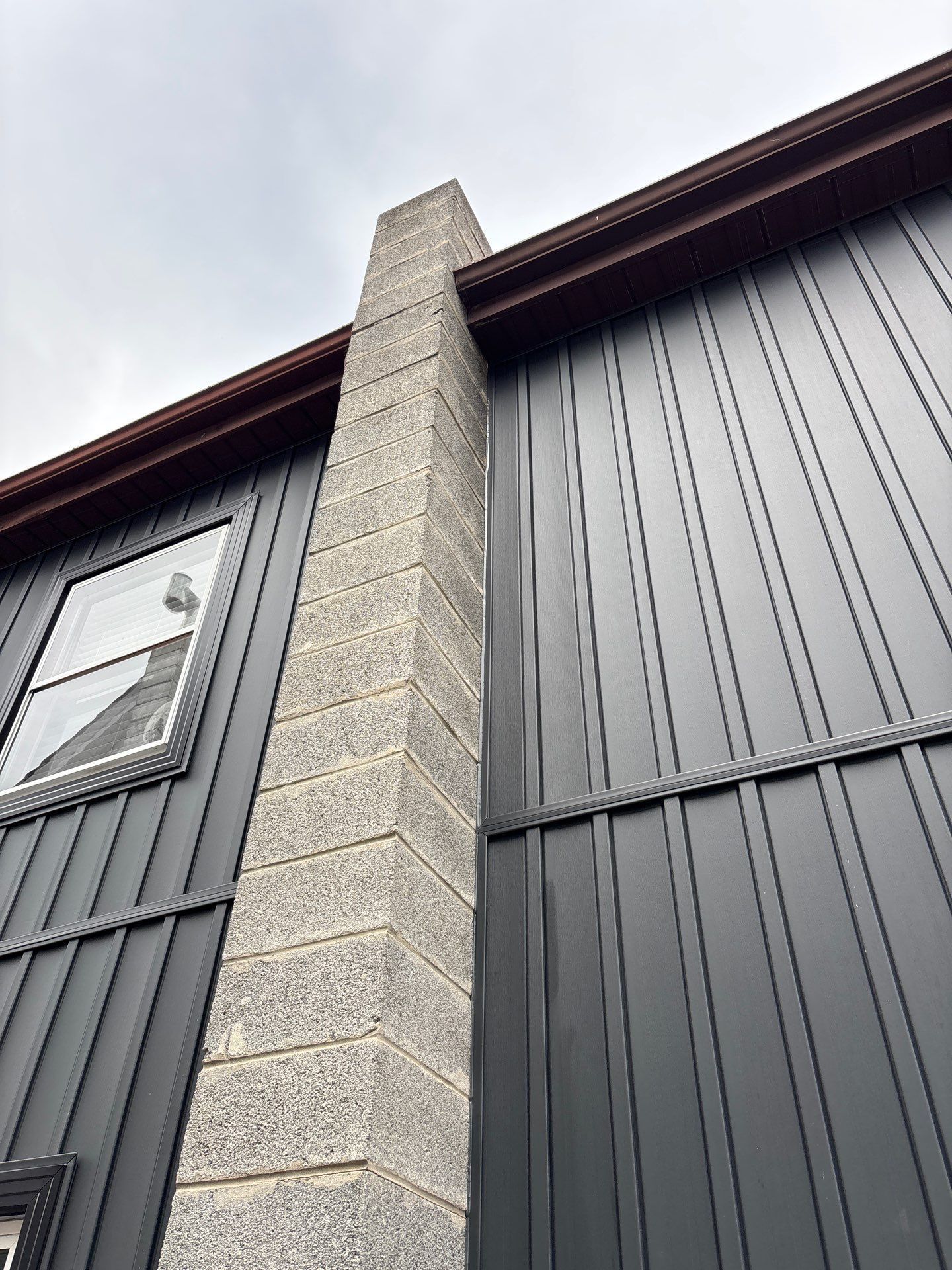 Stone chimney against dark gray building with vertical siding and brown trim. Cloudy sky.