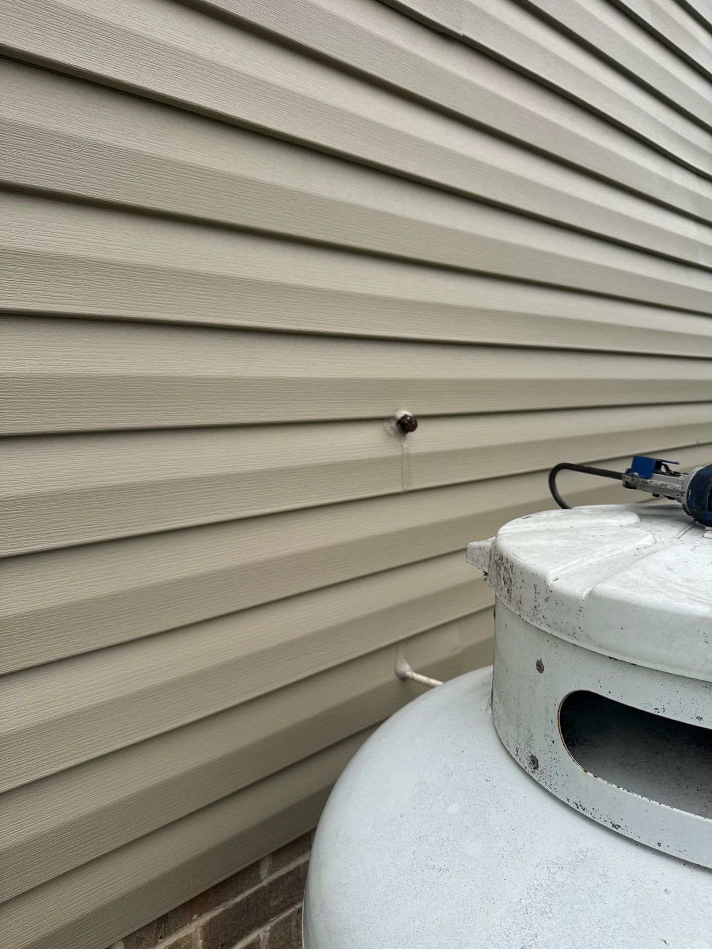 Tan vinyl siding with a black object mounted, next to a white propane tank.