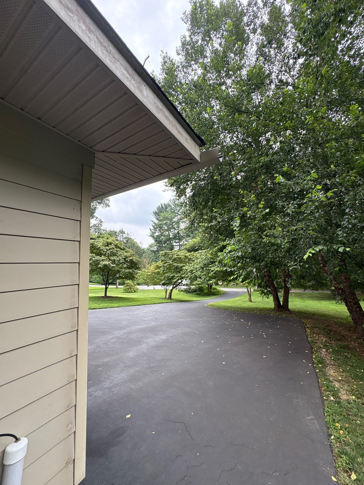 Asphalt driveway beside a building with beige siding, trees, and a cloudy sky.