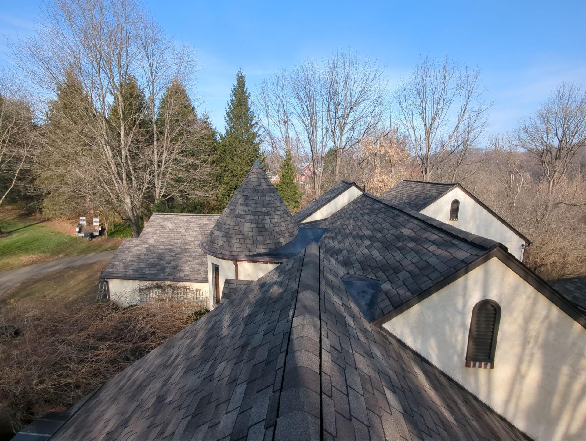 Overhead view of a house with multiple roof peaks; brown shingles and white walls; trees in the background.