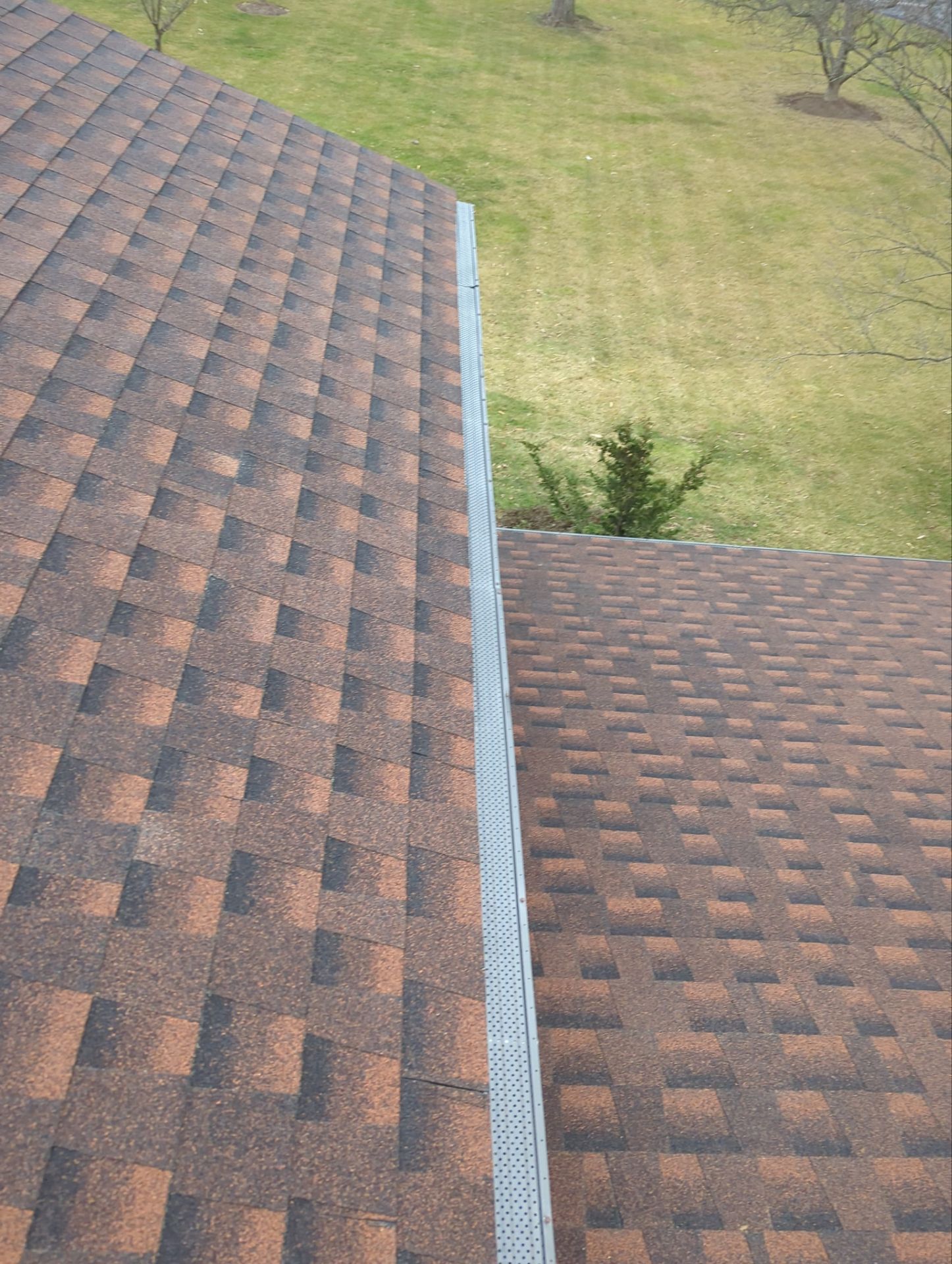 Two brown shingled roofs meet, a gray metal flashing between them, with a green yard beyond.