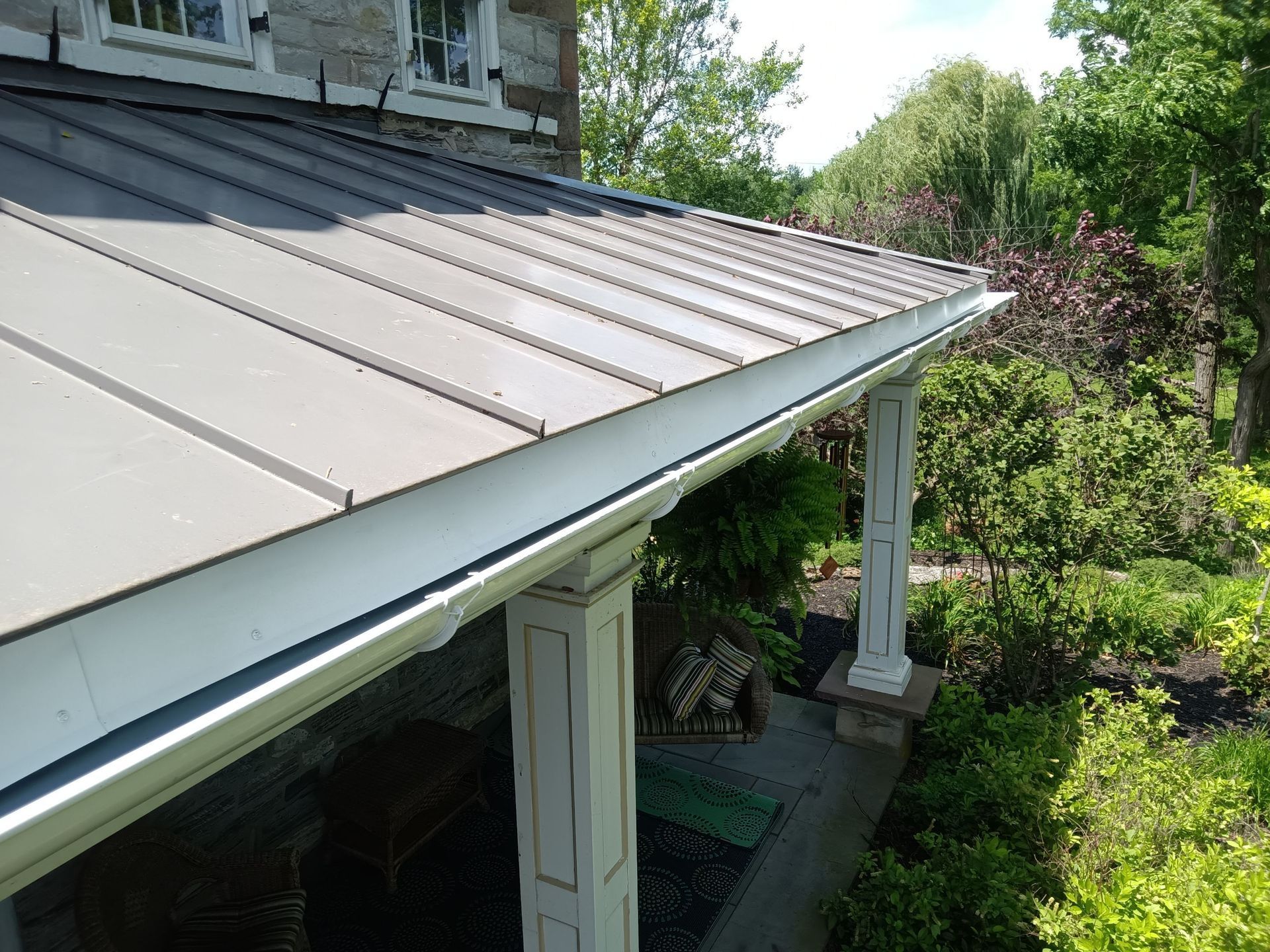 Two-story house with tan siding, black shutters, white porch with hanging plant, and a cloudy sky.