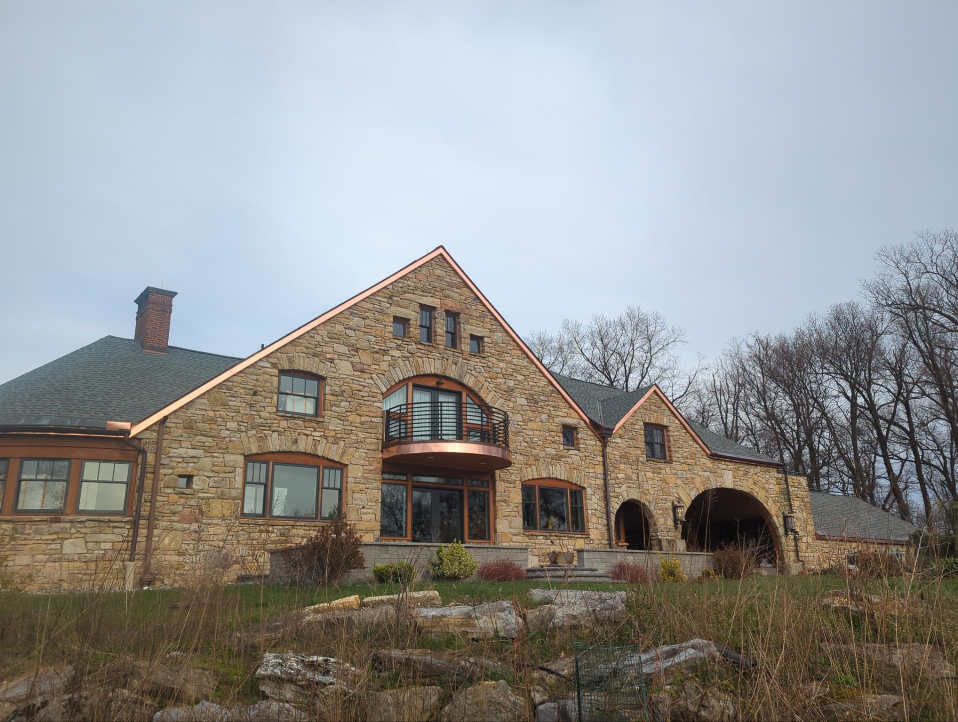 Stone house with copper trim and dark windows, set on a hillside under a cloudy sky.