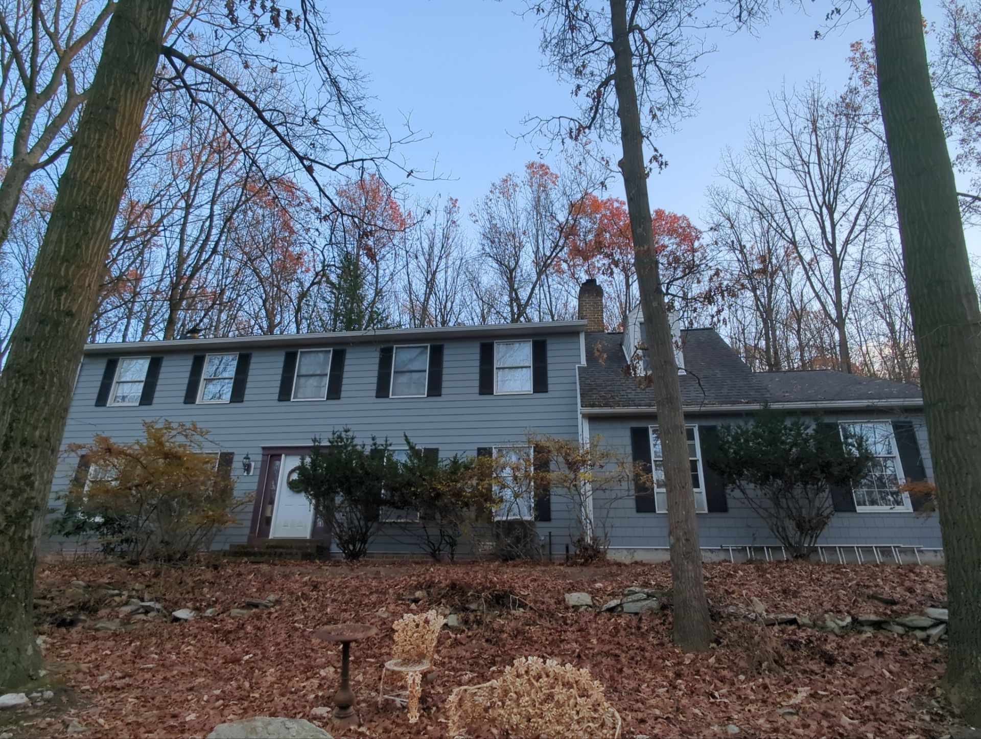 Two-story house with gray siding, black shutters, and a chimney surrounded by trees with fall foliage.