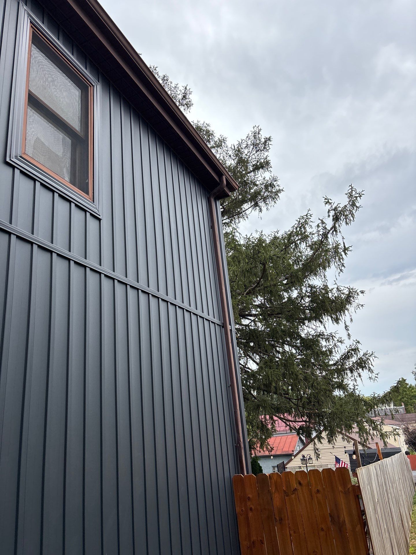 Dark grey house siding with a brown gutter and window, next to a tree and wooden fence.