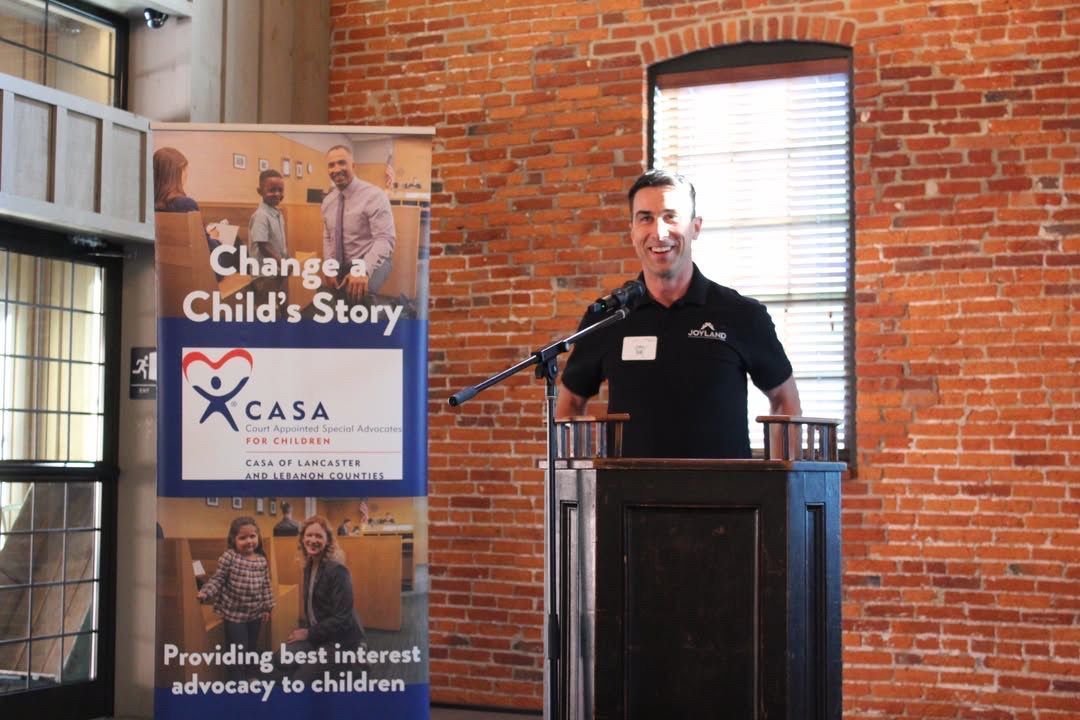 Man speaking at a podium next to a banner that says "Change Child's Story," in a room with a brick wall.