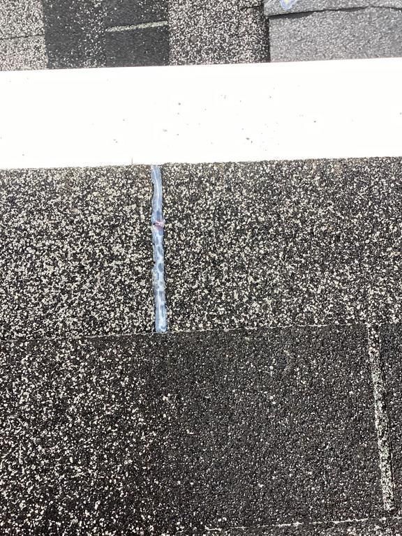 Close-up of asphalt shingles on a roof with a crack sealed with a light-colored sealant. White trim board visible.
