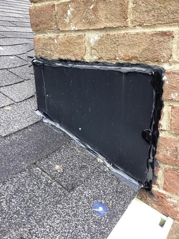 Black flashing sealed with caulk meets brick chimney on a shingled roof.