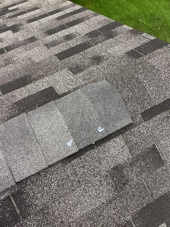 Close-up of a gray asphalt shingle roof. One shingle is lifted, with two nails visible. Green grass in the background.