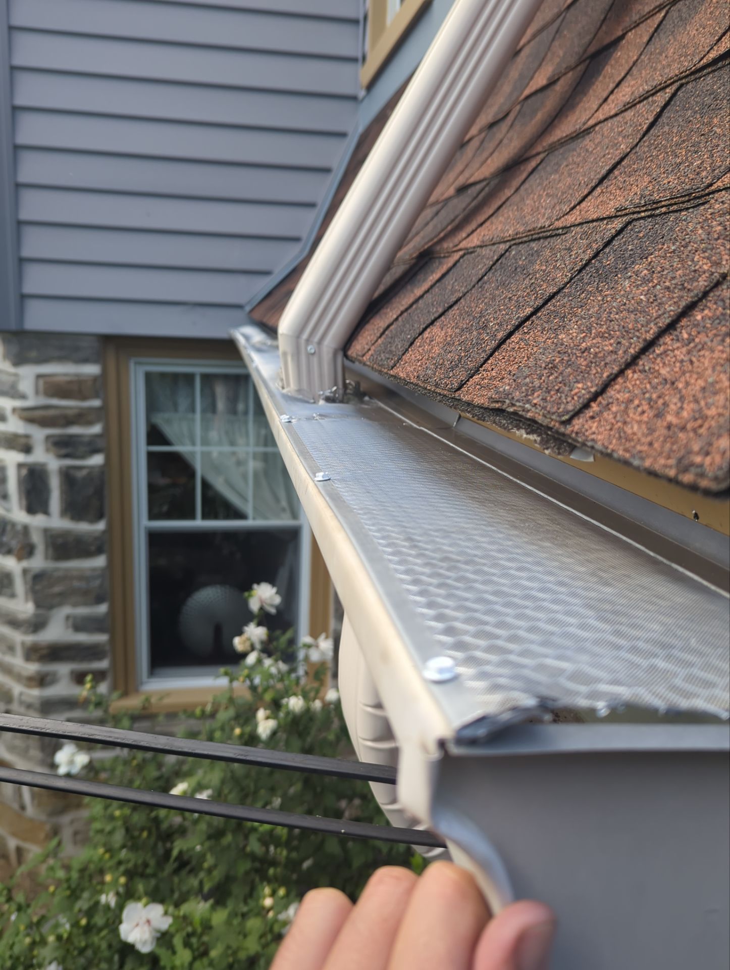 Two-story house with tan siding, black shutters, white porch with hanging plant, and a cloudy sky.