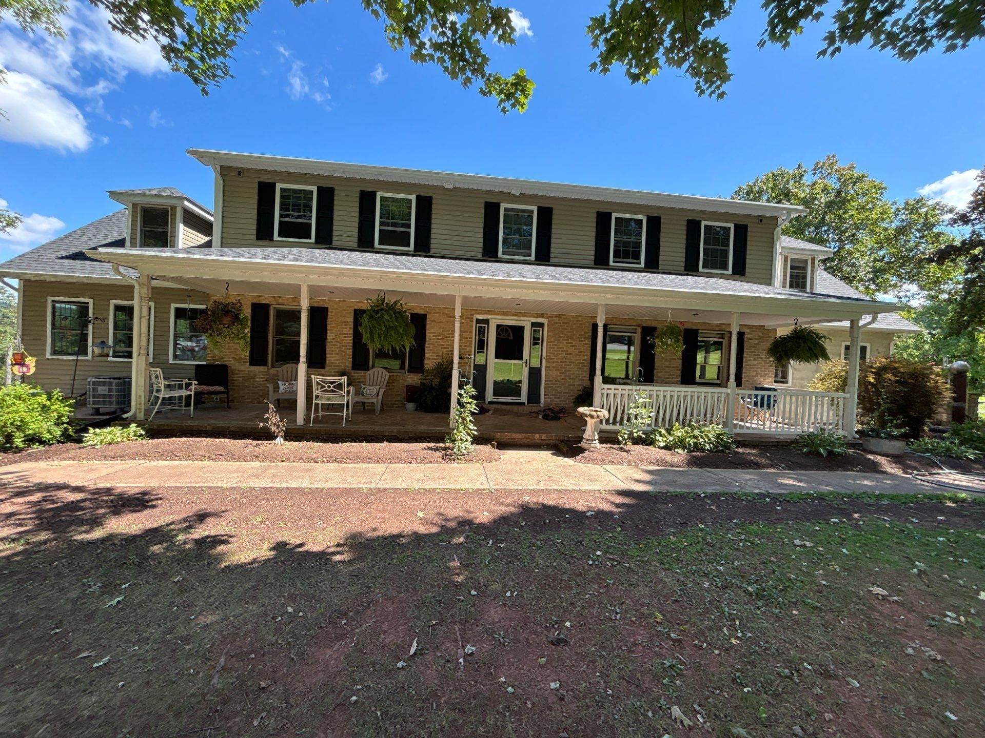 Two-story beige house with black shutters and a large front porch under a sunny blue sky.