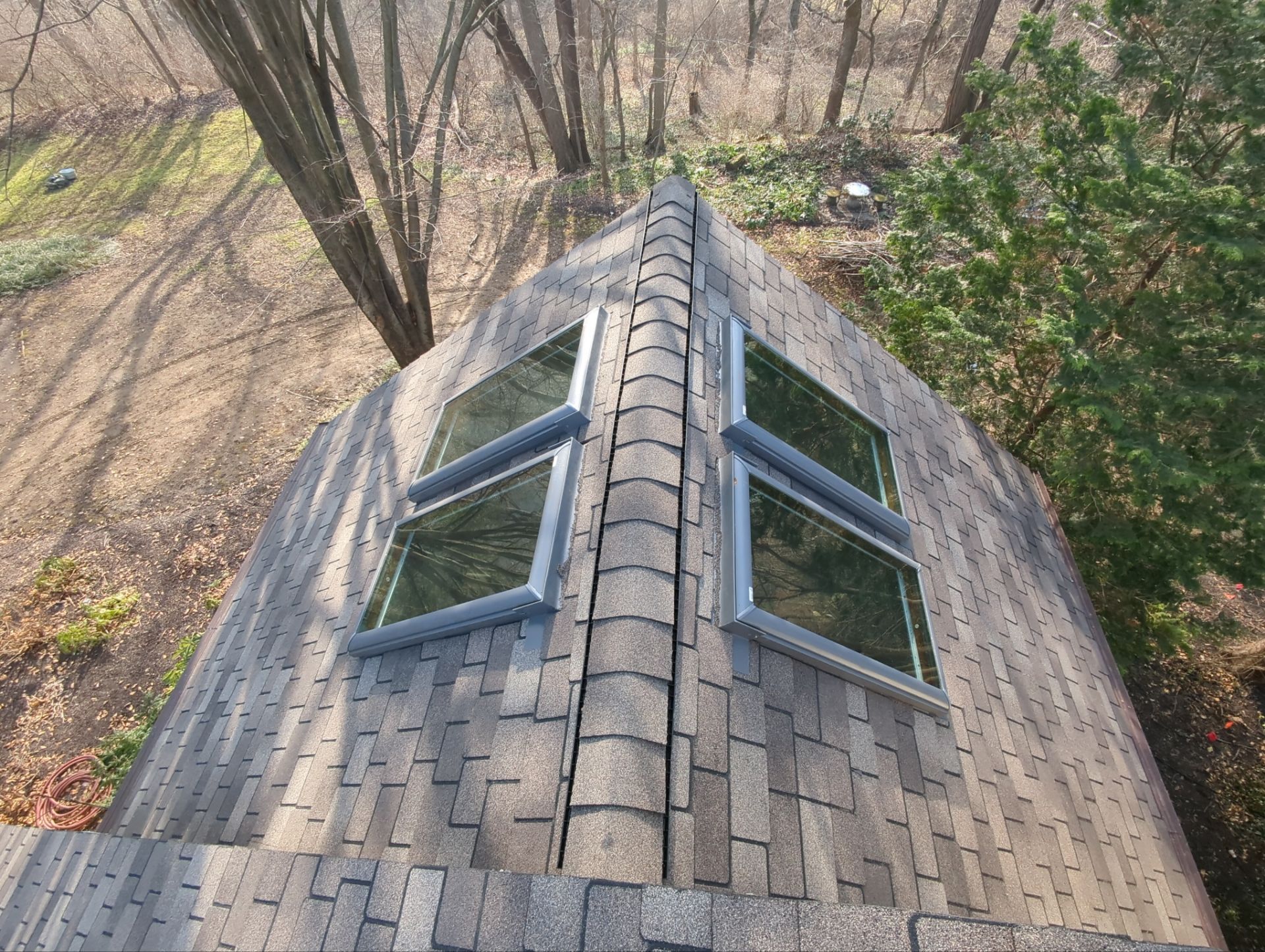 Overhead view of a roof with shingles, ridge cap, and four skylights surrounded by trees.