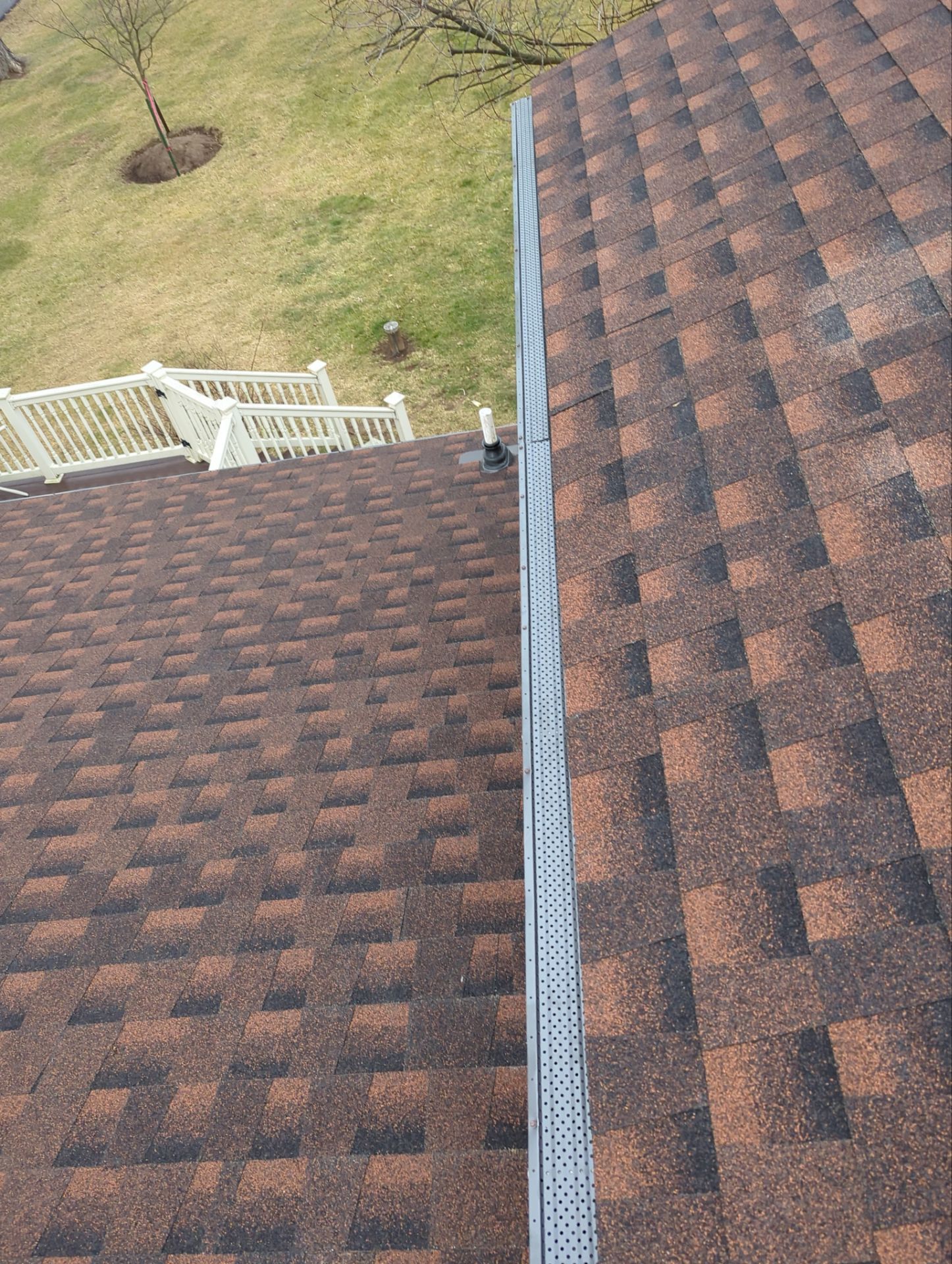 Two brown shingled roofs meet with a gray gutter guard on a sunny day. A white fence is visible.