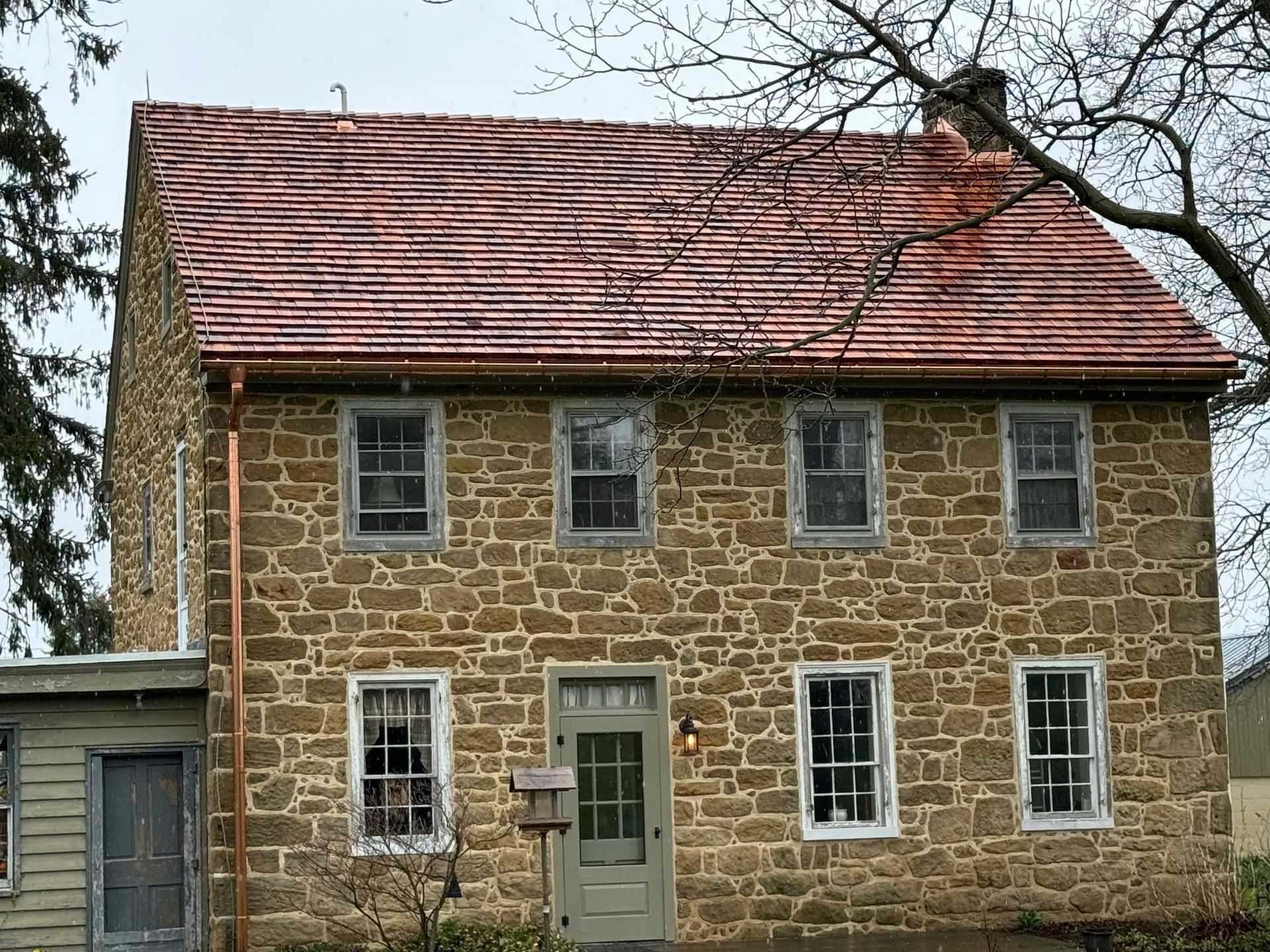 Two-story stone house with red tile roof, six windows on front, a side addition, and leafless tree.