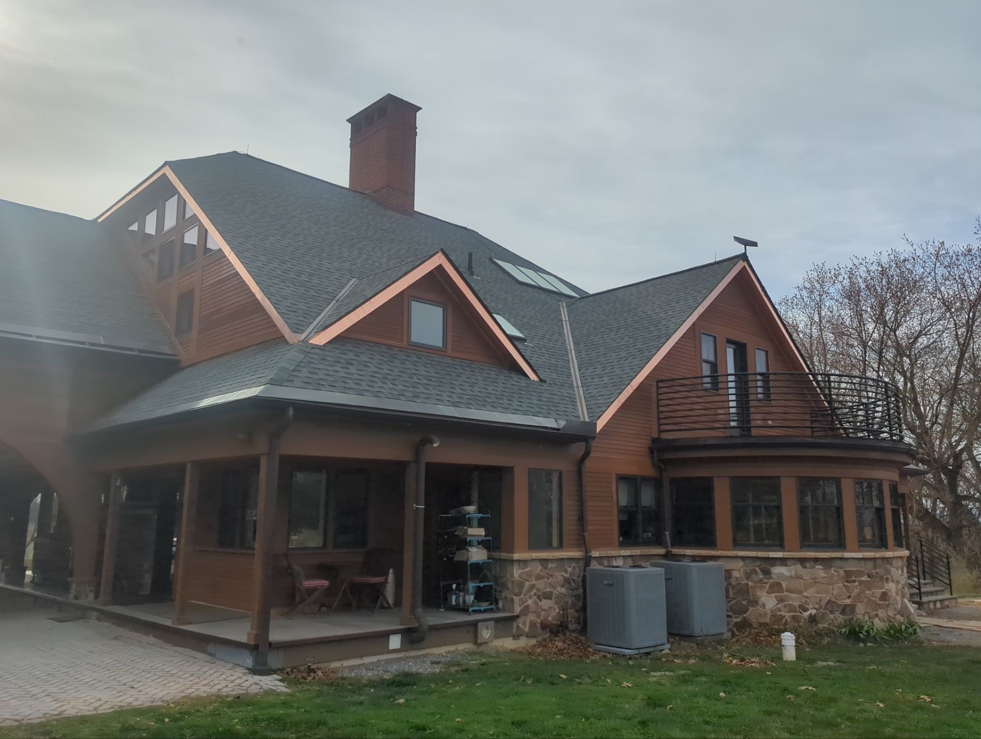 Large brown house with gray shingled roof, copper trim, stone base, and porch. Chimney and small balcony visible.