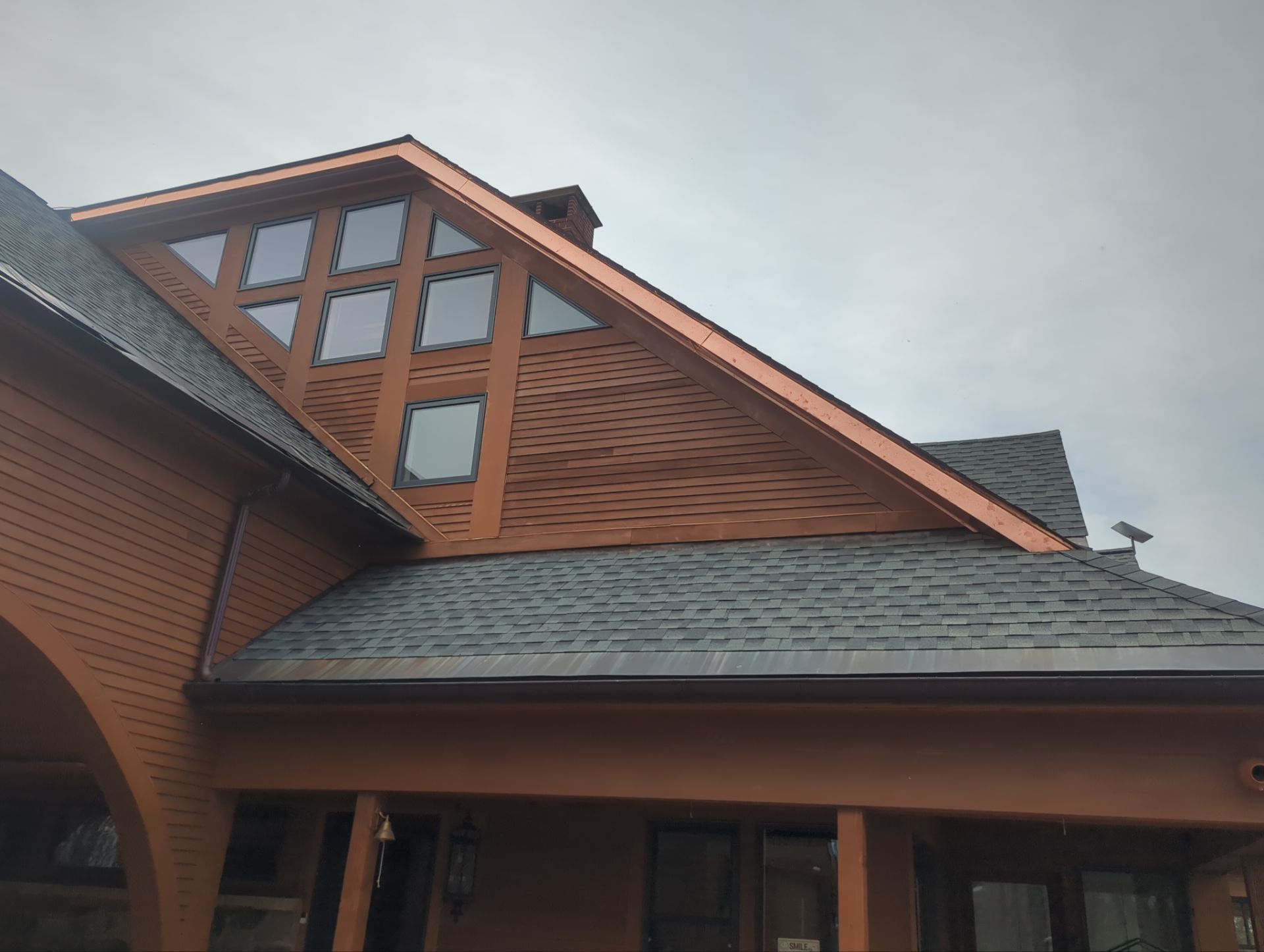 Brown roof with triangular window section and dark gray shingles, under cloudy sky.