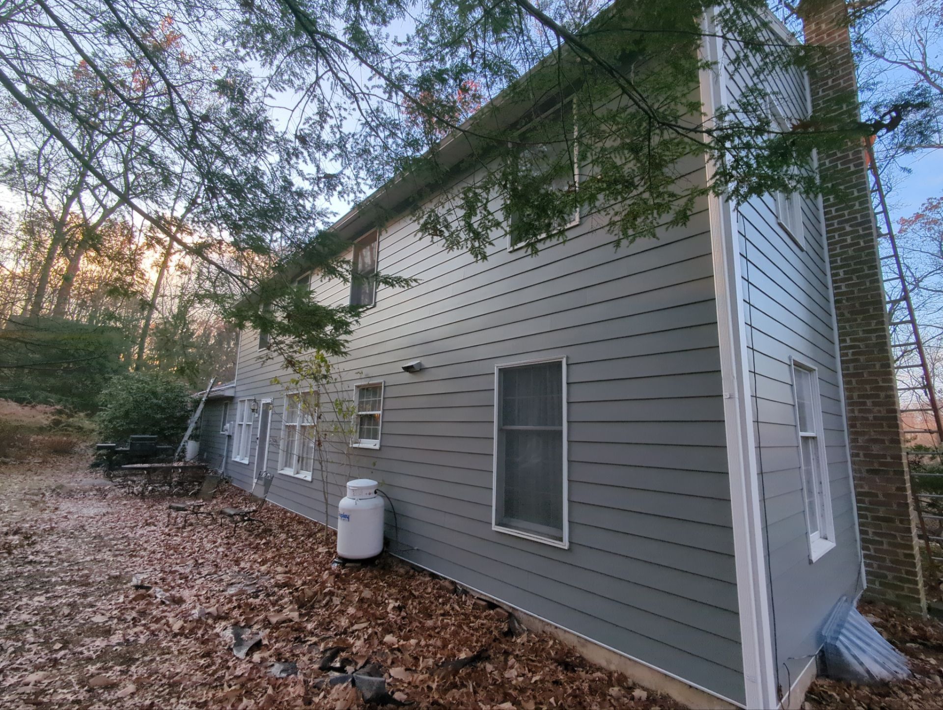 Side view of a two-story gray house with a propane tank, surrounded by fall foliage and trees.