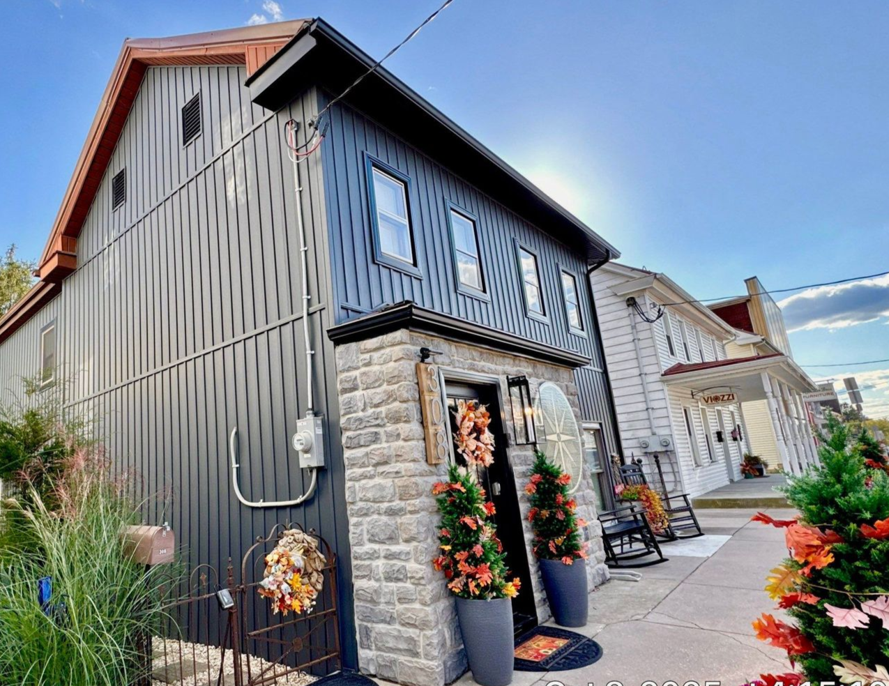Two-story house with tan siding, black shutters, white porch with hanging plant, and a cloudy sky.