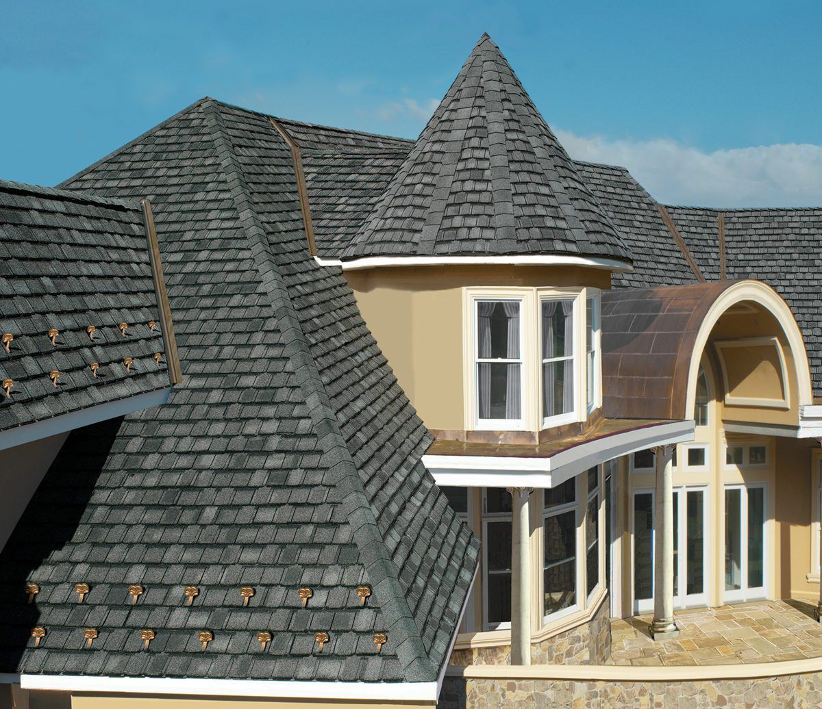 Gray shingled roof of a house with a turret and curved architectural elements. Sunny day.
