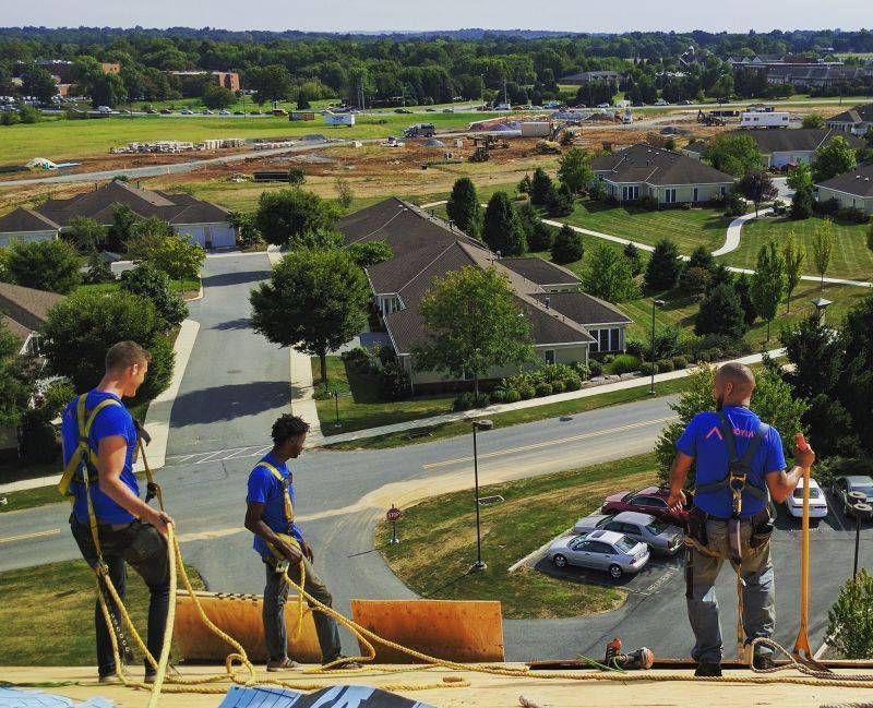 Three roofers in blue shirts and safety harnesses work on a roof overlooking a suburban neighborhood.