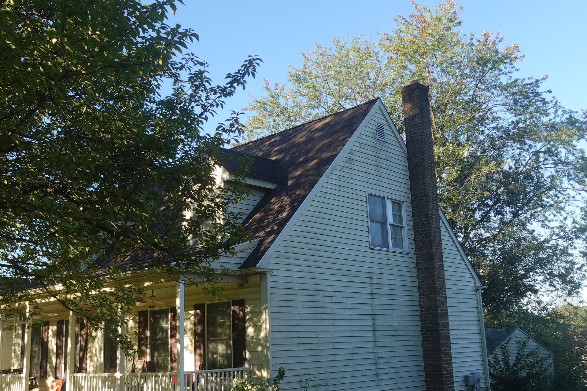 Two-story, light-colored house with dark roof and tall brick chimney, partially obscured by green trees.