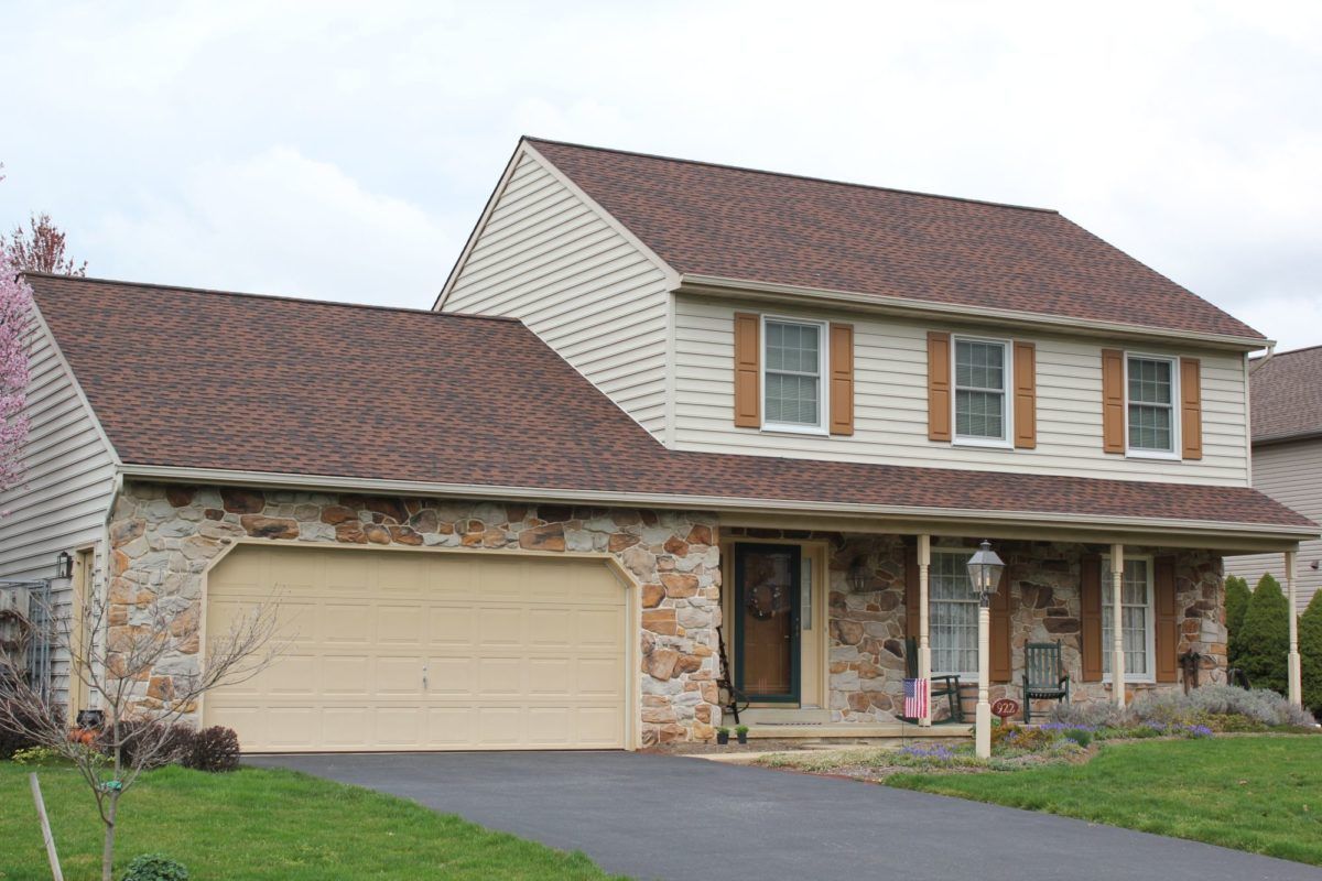 Two-story house with stone facade and tan siding. Brown roof, beige garage door, and green lawn.