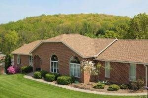 Brick ranch house with a brown roof and a green lawn, set against a hill covered in trees.
