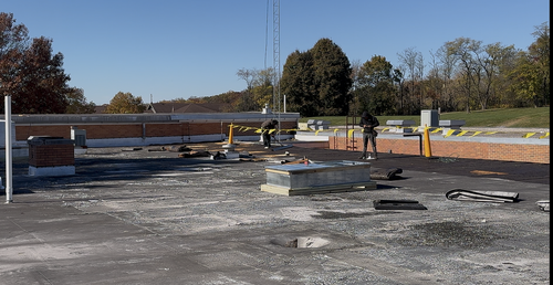 Construction workers on a roof surrounded by debris; clear sky and trees in the background.