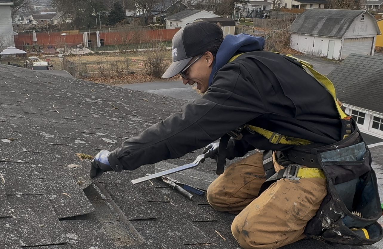 Roofer kneels on a weathered roof, smiling while using a tool. Wearing a harness and tool belt.
