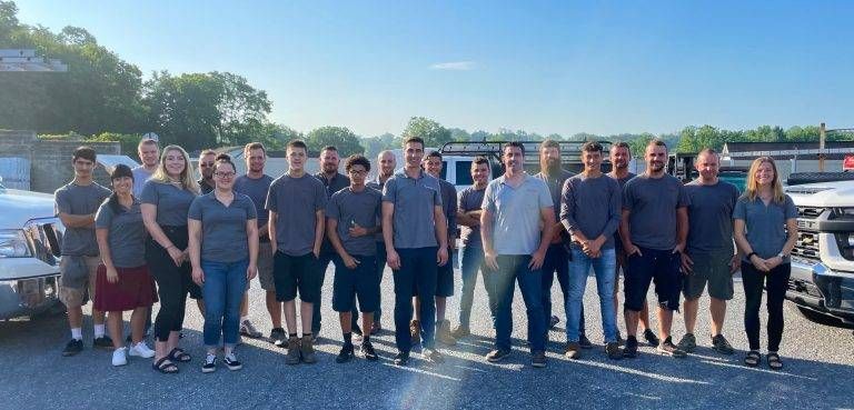 Group of people standing outdoors, all in gray shirts. Blue sky and some vehicles in the background.