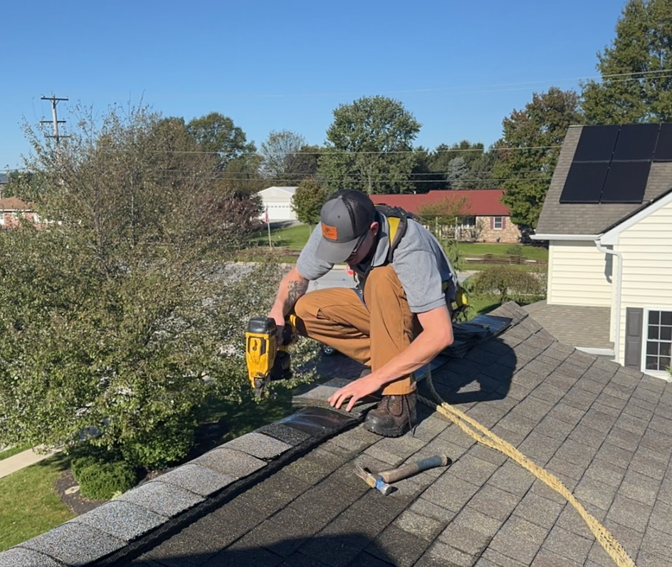 Man on a ladder installing a white gutter on a house, sunny outdoor setting.