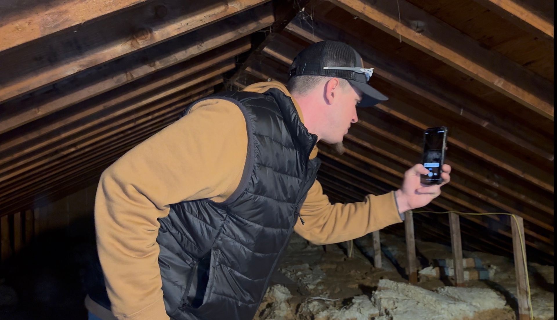 A person wearing a hat and quilted vest stands in an attic, holding up a smartphone to record or photograph the space.