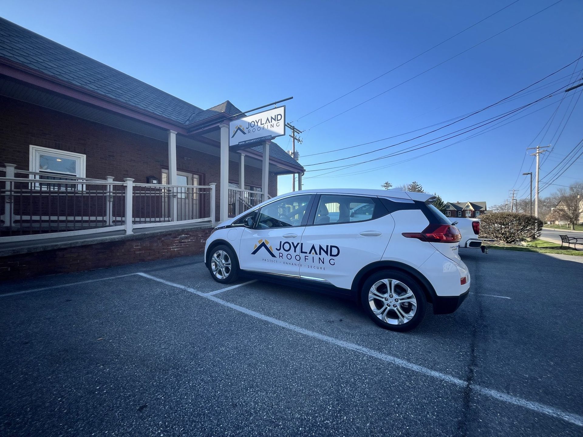 White electric car with company logo parked outside a building with the same logo. Blue sky.